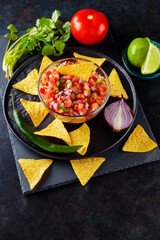 Mexican Pico de Gallo salsa and ingredients. Bowl of tasty Pico de Gallo salsa and nachos on dark background. Top view. Copy space