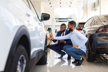 Young Black Man Choosing New Car In Dealership Center, Examining Tires