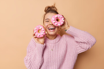 Happy joyful woman covers eye with delicious glazed doughnut laughs happily dressed in casual...