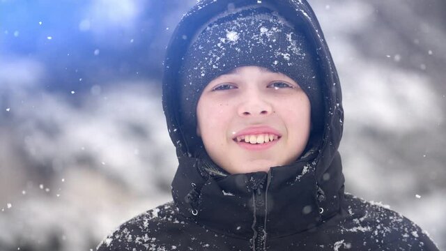 Portrait of a European teen boy in snowfall in winter, he stands on the street and looks at the camera and looks happy. Slow motion, shallow depth of field
