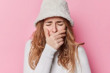 Upset crying young woman covers mouth with hand feels displeased has problems being in despair stands distressed wears winter hat and jumper isolated over pink background. Emotional burnout.