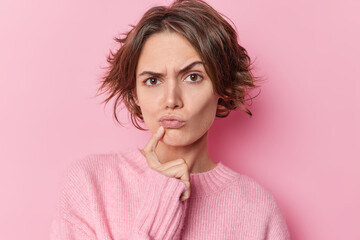 Serious skeptical woman keeps index finger near folded lips raises eyebrows suspiciously has doubts suspects someone tries to search solution wears casual jumper isolated over pink background