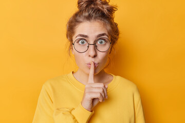 Stunned emotional beautiful woman hushing with index finger shares secret makes taboo gesture stares through round spectacles wears casual jumper isolated over yellow background. Shh be quiet - obrazy, fototapety, plakaty