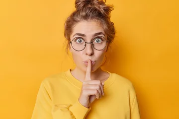 Foto op Canvas Stunned emotional beautiful woman hushing with index finger shares secret makes taboo gesture stares through round spectacles wears casual jumper isolated over yellow background. Shh be quiet © Wayhome Studio