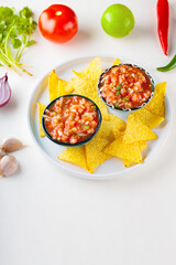 Salsa sauce and nacho chips on white plate. Mexican homemade salsa dip and ingredients on a white background. Copy space. Top view
