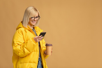 Modern zoomer blogger. Excited woman in jacket and glasses holding takeaway coffee and reading message on smartphone