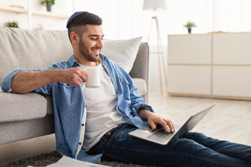 Smiling jewish man watching video on computer, drinking coffee