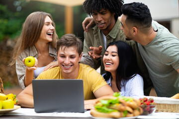 Group of diverse young friends using laptop while camping in countryside, communicating online, having fun