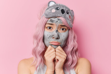 Upset discontent woman with dyed hair keeps hands under chin feels sad applies beauty mask for skin refreshment undergoes face care procedures before sleeping time isolated over pink background