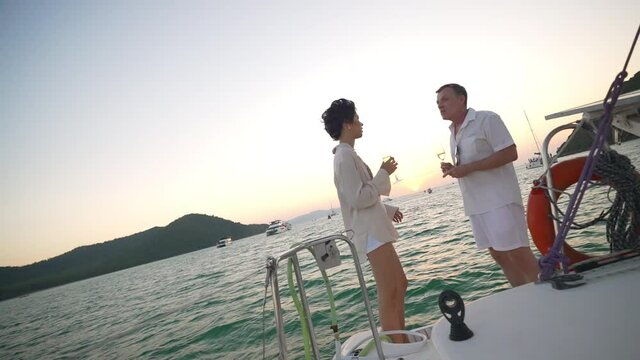 4K Caucasian couple enjoy outdoor luxury party drinking champagne with talking together while catamaran boat sailing at sunset. Man and woman relax with outdoor lifestyle on summer travel vacation