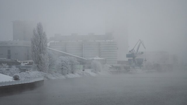 Bremen, Germany. First hoarfrost at Weserwehr weir next to swb Stadtwerke power plant beneath Weser river in foggy ice cold Hastedt. Chemical industry and moody bridges. luffing crane in front.