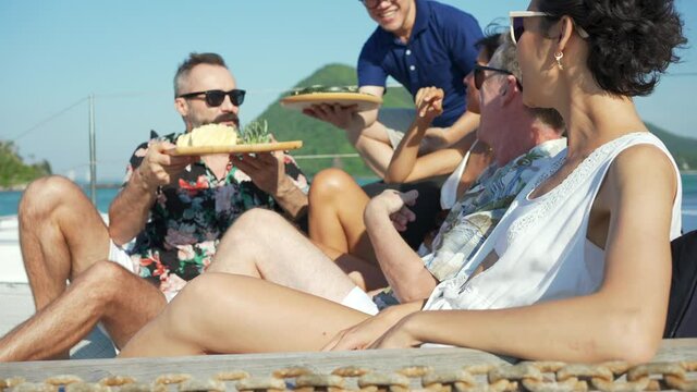 4K Group of Caucasian people friends enjoy luxury outdoor party eating fresh fruit together while catamaran boat sailing. Man and woman relaxing outdoor lifestyle sail yacht on summer travel vacation