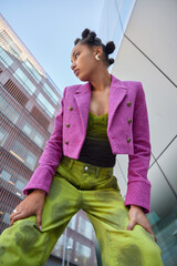 Vertical shot of stylish woman dressed in pink jacket and green trouers leans on knees focused away...