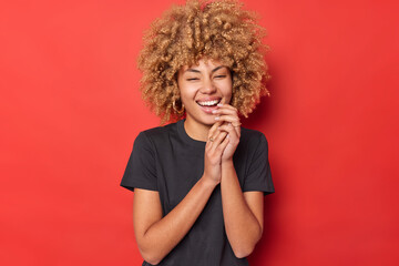 Beautiful curly haired woman keeps hands together laughs out happily smiles toothily wears casual black t shirt hears something funny wears casual black t shirt isolated over vivid red background