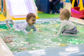 two little cute baby toddler girls in a dress sit, lie and laugh enjoying in a toy pool on a...