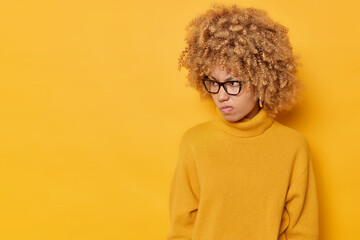 Fototapeta na wymiar Curly haired young cute woman looks angrily away being dissatisfied with something purses lips concentrated on left wears eyeglasses and jumper isolated over yellow background with copy space