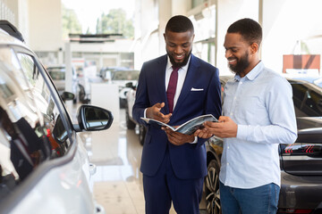 Black Male Customer In Car Dealership Center Looking At Brochure With Salesman