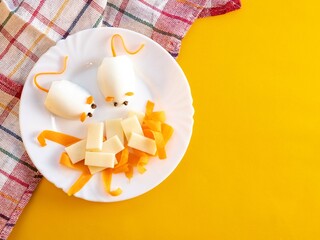 Children's food from boiled mouse eggs with carrot ears, cheese slices in a white plate on a yellow background. Food for a children's party. Place of text.
