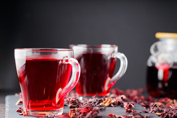 Hibiscus tea in glass mug on a dark background. Cup of red hibiscus tea and dry hibiscus petals on a slate board