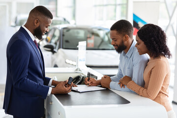 Buying New Car. African American Couple Signing Papers In Dealership Center