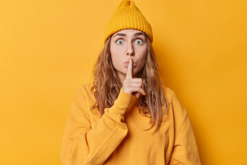 Surprised stunned young woman looks concerned makes silence gesture demands to be quiet wears hat...