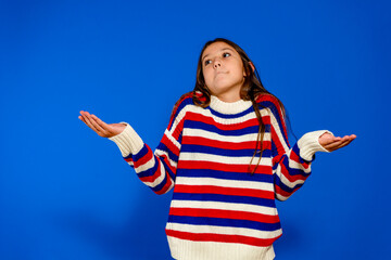 Confused and doubtful cute little girl dressed in a striped sweater raising her hands to keep a copy space. Isolated on blue studio background