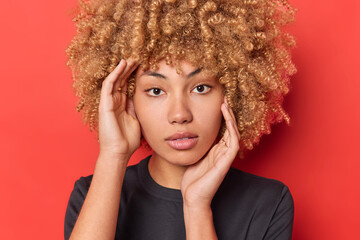 Close up portrait of self confident serious woman with curly blonde hair touches face looks directly at camera wears casual black t shirt isolated on red background shows natural beauty without makeup