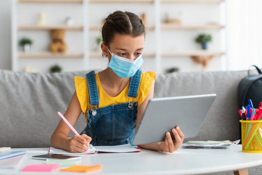 Schoolgirl in medical mask studying at home with digital tablet