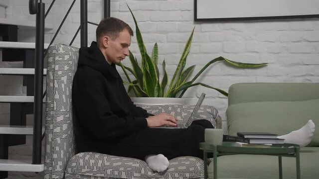 a young guy sits near the stairs in the apartment and types something on his laptop