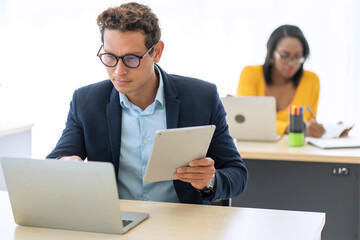 Handsome male finance worker sitting in a conference room holding a tablet and working on a laptop giving financial advice at his office desk.