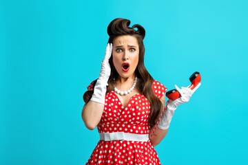 Terrified millennial woman in retro pinup dress holding landline phone receiver, touching her temples on blue background