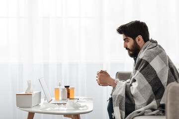 Sick at home. Young arab man having fever and drinking hot tea, sitting on sofa and watching film on laptop, side view