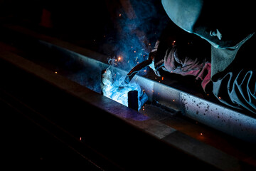 A man wearing a welding mask welds metal parts at a factory.