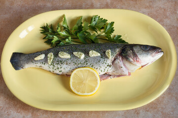 Overhead view of a European seabass garnished with chopped garlic and spices in a single file on its raw skin accompanied by sprigs of fresh parsley and a lemon slice on a colorful ceramic platter on