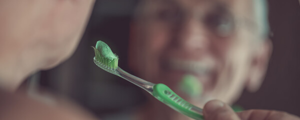 Green toothpaste on toothbrush in hand of senior man looking in mirror - obrazy, fototapety, plakaty