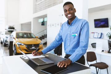 Fototapeta Portrait of happy car salesman standing at work desk, smiling at camera, using laptop and touch pad in auto dealership obraz