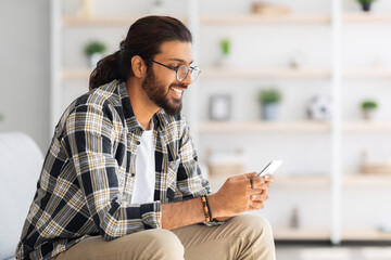 Positive long-haired middle-eastern guy sitting on couch, using cellphone