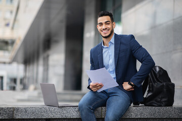 Handsome arab entrepreneur sitting by office building with laptop