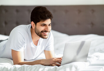 Dating web site, free time with device. Happy young arab man lying on bed and typing on laptop