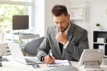 Naklejka premium Thoughtful young businessman writing on clipboard at table in office