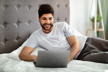 Online work, study, courses, new normal and be safe during COVID-19. Happy arab man lying on bed, looking at laptop