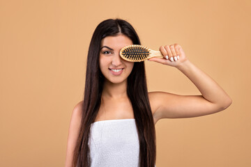 Hair care concept. Happy armenian young woman with long fair hair covering eye with hair brush, beige background