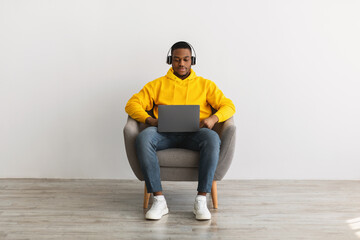 Serious African Guy Using Laptop Wearing Headphones Over Gray Wall