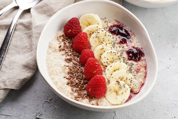 Trendy breakfast bowl with oatmeal porridge, fresh raspberries, banana, chia seeds, linseed and jam