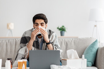 Sick arab man sneezing nose in paper tissue and looking at laptop screen, sitting at home and working, empty space - obrazy, fototapety, plakaty