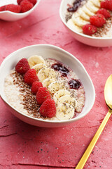 trendy breakfast bowl with oatmeal porridge, fresh raspberries, banana, chia seeds, linseed and jam on magenta surface