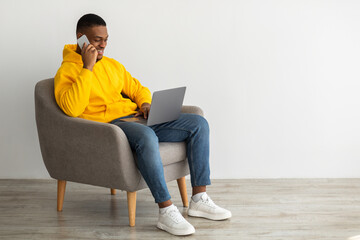 African Guy Talking On Cellphone Using Laptop Over Gray Wall