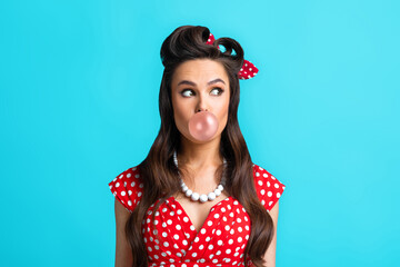 Stylish young pin up lady in retro outfit blowing bubble from chewing gum, looking aside over blue studio background