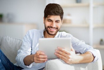 Technology concept. Portrait of happy arab man using digital tablet sitting on the couch at his...