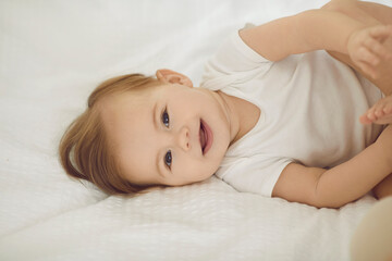 Baby in white bedding. Close up portrait of smiling healthy beautiful six month old baby girl lying on light bed. Side view of adorable baby in white bodysuit lying and playfully looking at camera.
